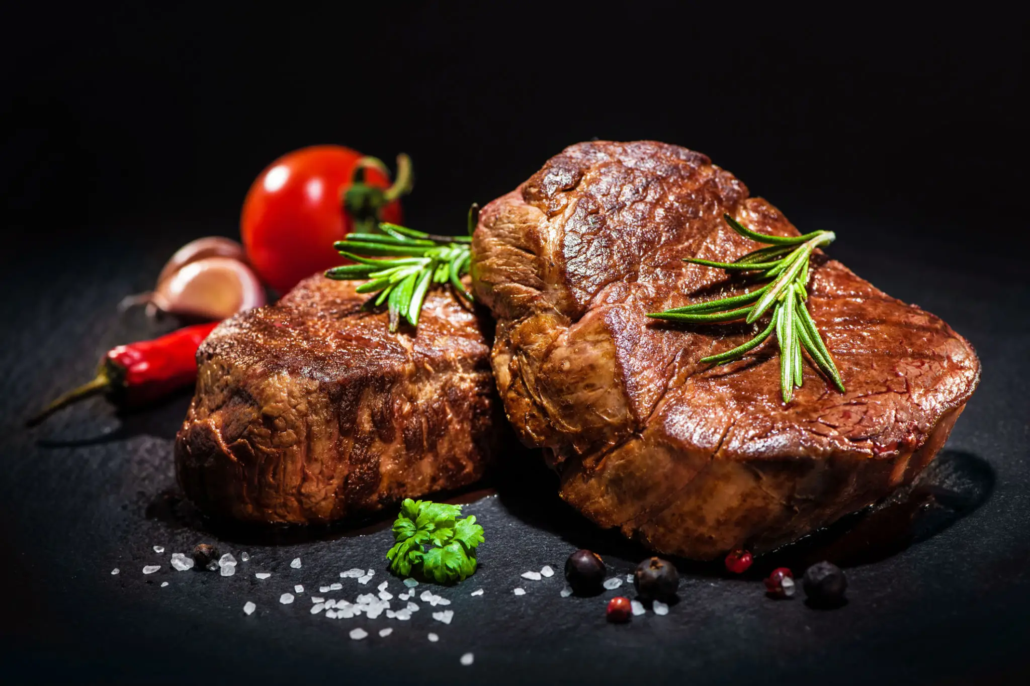 Two grilled steaks garnished with rosemary and spices on a dark surface.